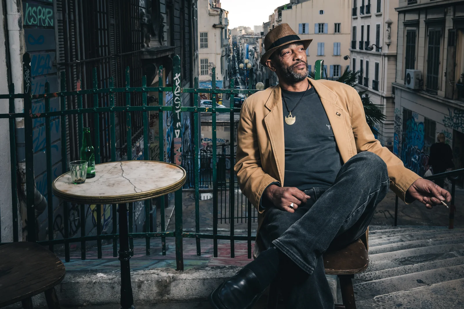 Man in a tan jacket and hat seated beside a café table.