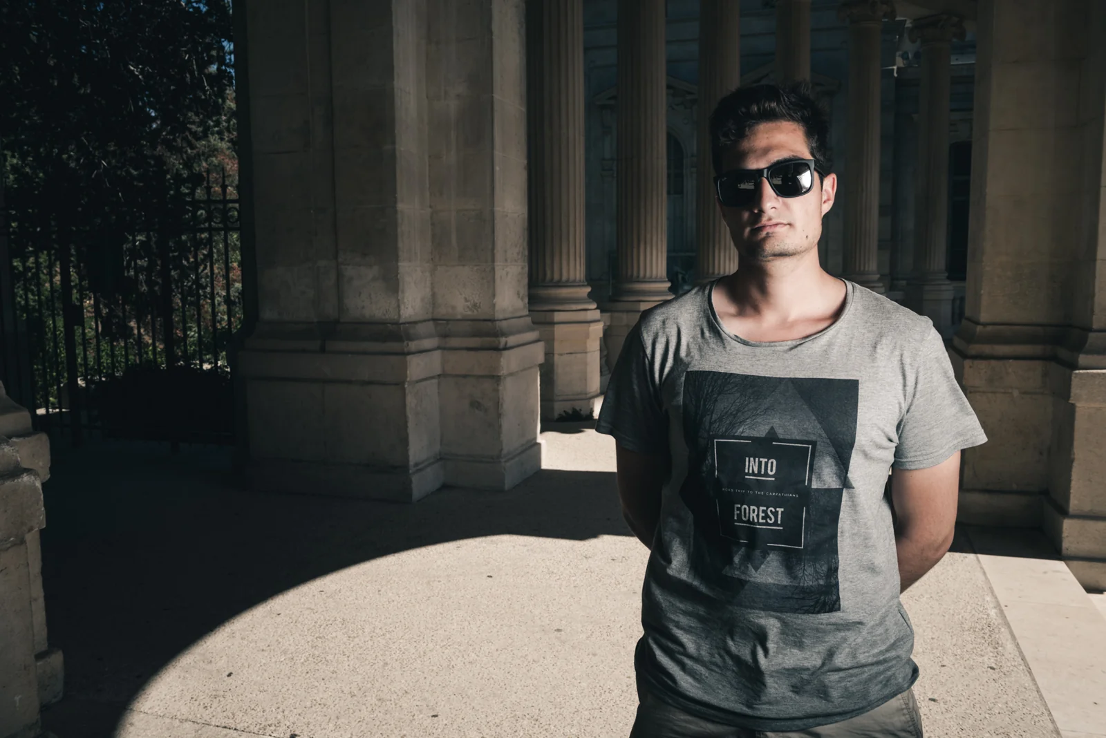 Man in sunglasses standing in the shade of a stone colonnade.